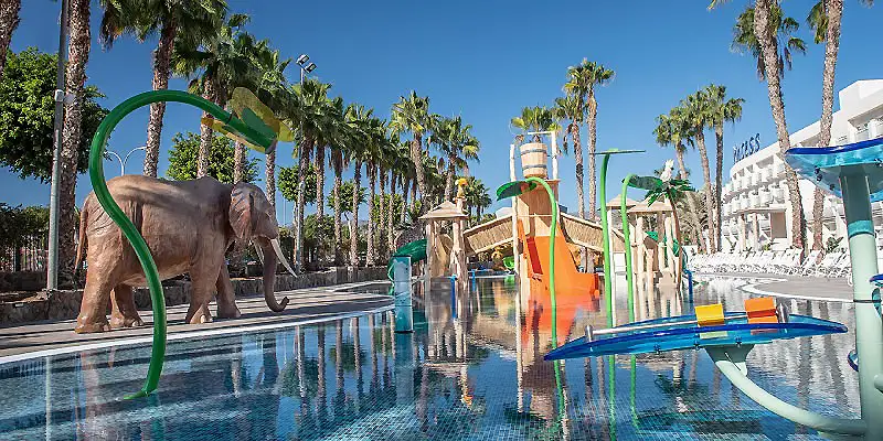Elefante en piscina de parque acu&aacute;tico con tobog&aacute;n y palmeras al fondo