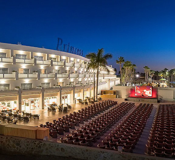 Auditorio con filas de sillas, hotel iluminado y palmeras al anochecer