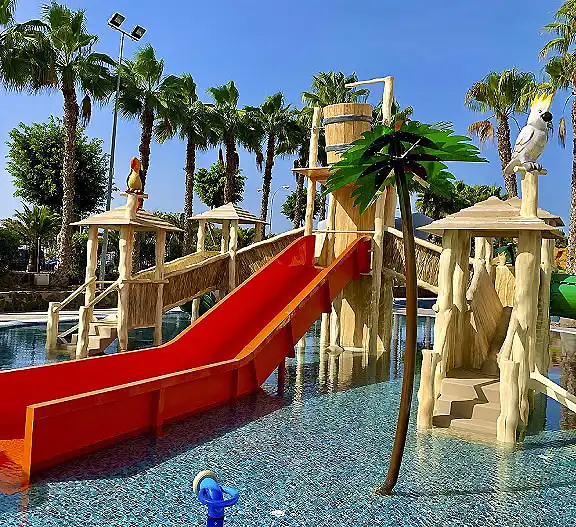 Tobog&aacute;n rojo en piscina infantil con palmeras y figuras de madera.