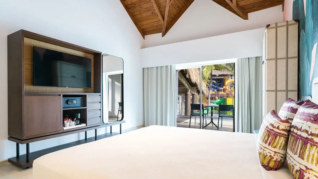 Habitaci&oacute;n moderna con cama, TV y cortinas, vista a piscina y palmeras.