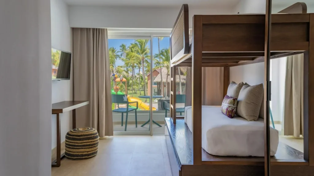 Habitaci&oacute;n de hotel con cama, cortinas y vista a palmeras y piscina.