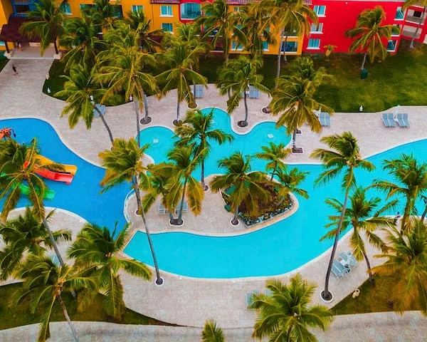 Piscina azul rodeada de palmeras y edificio multicolor, vista a&eacute;rea.