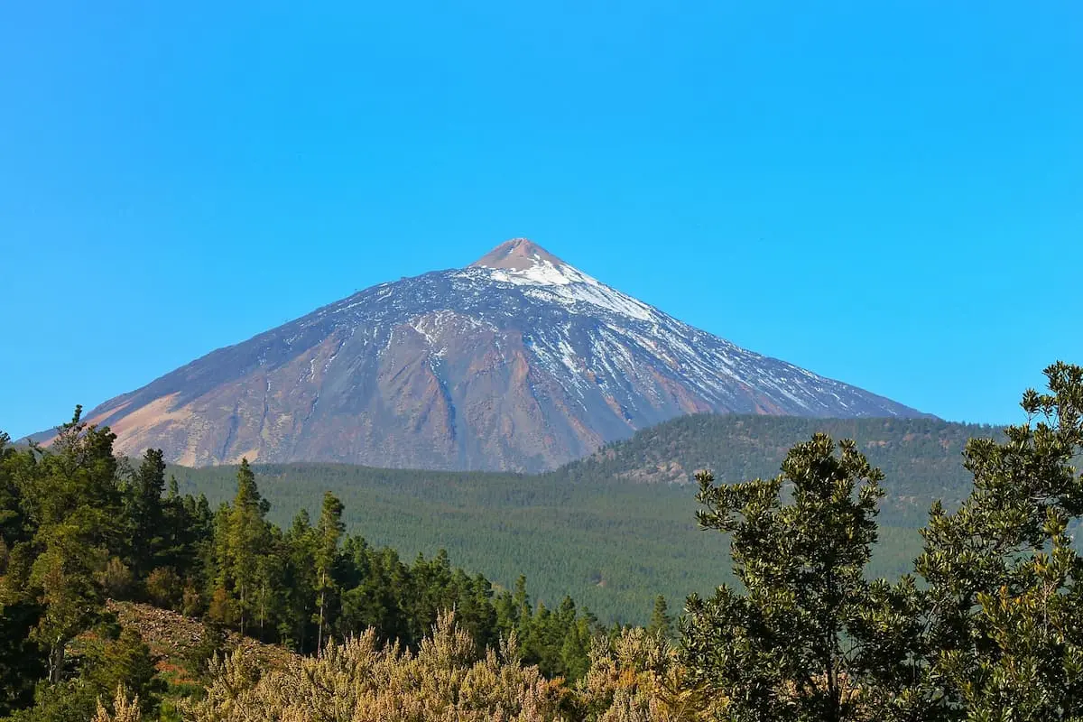 Monta&ntilde;a volc&aacute;nica con nieve en la cima, rodeada de bosques y cielo azul