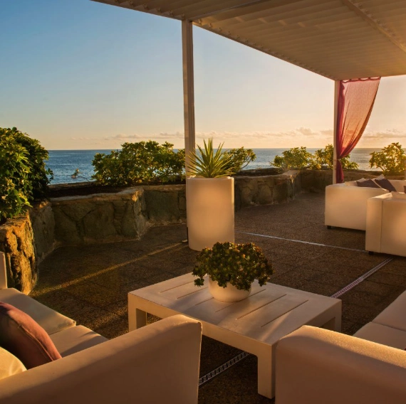Terraza con sof&aacute;s y vista al mar al atardecer, bajo toldo.