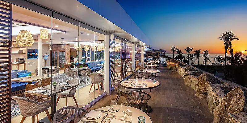 Terraza de hotel al atardecer con mesas, palmeras y vistas al mar.