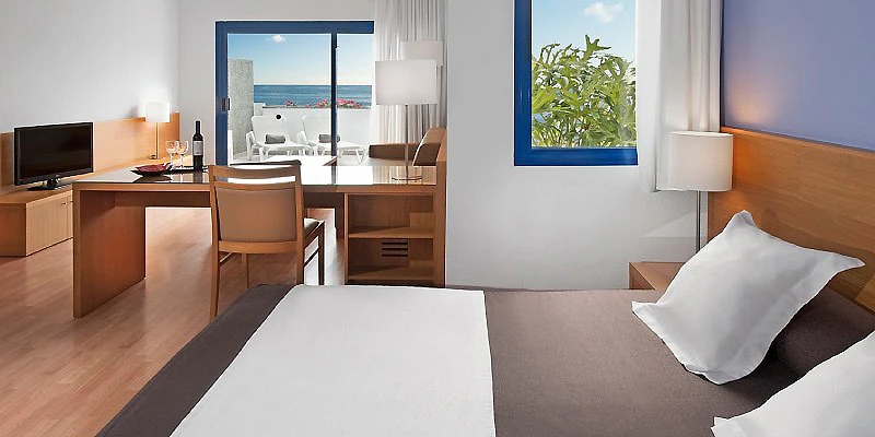 Habitaci&oacute;n de hotel con cama blanca, escritorio y vista al mar.