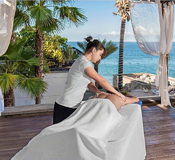 Masaje a una mujer sobre cama blanca frente al mar, con palmeras y terraza.