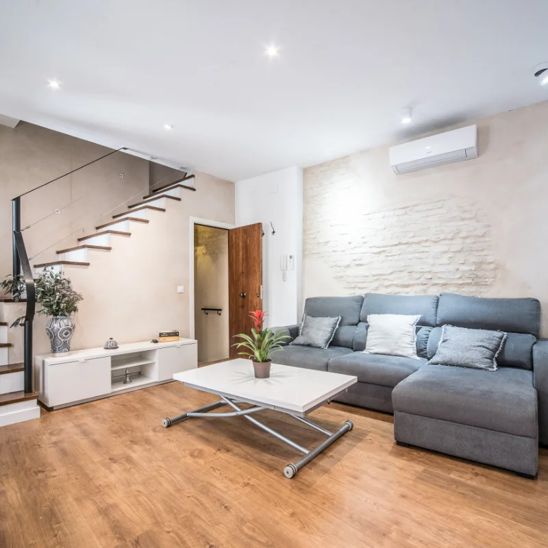 Sala moderna con sof&aacute; gris, mesa blanca y escalera de dise&ntilde;o minimalista.