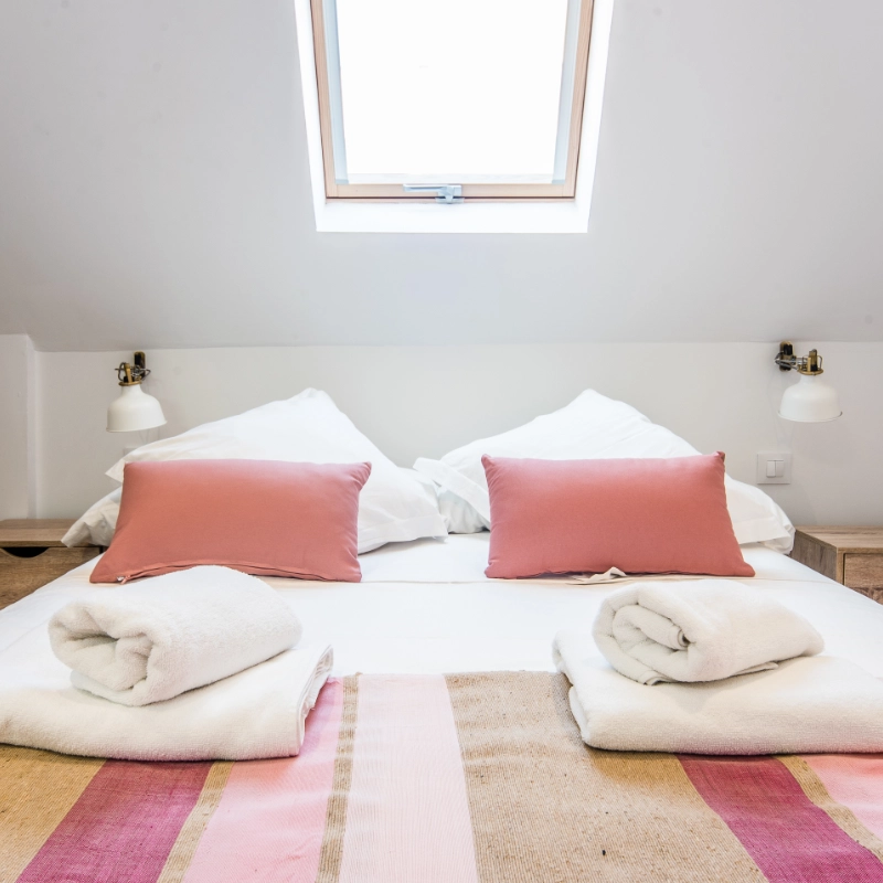 Cama con cojines rosas y manta a rayas bajo una ventana inclinada.