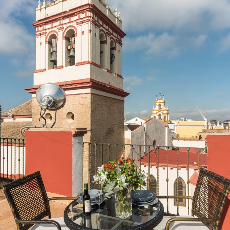 Terraza con mesa, flores y vistas a campanarios bajo un cielo despejado.