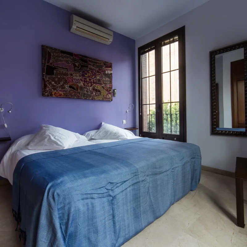Habitaci&oacute;n con cama doble, pared morada, espejo y ventana grande.