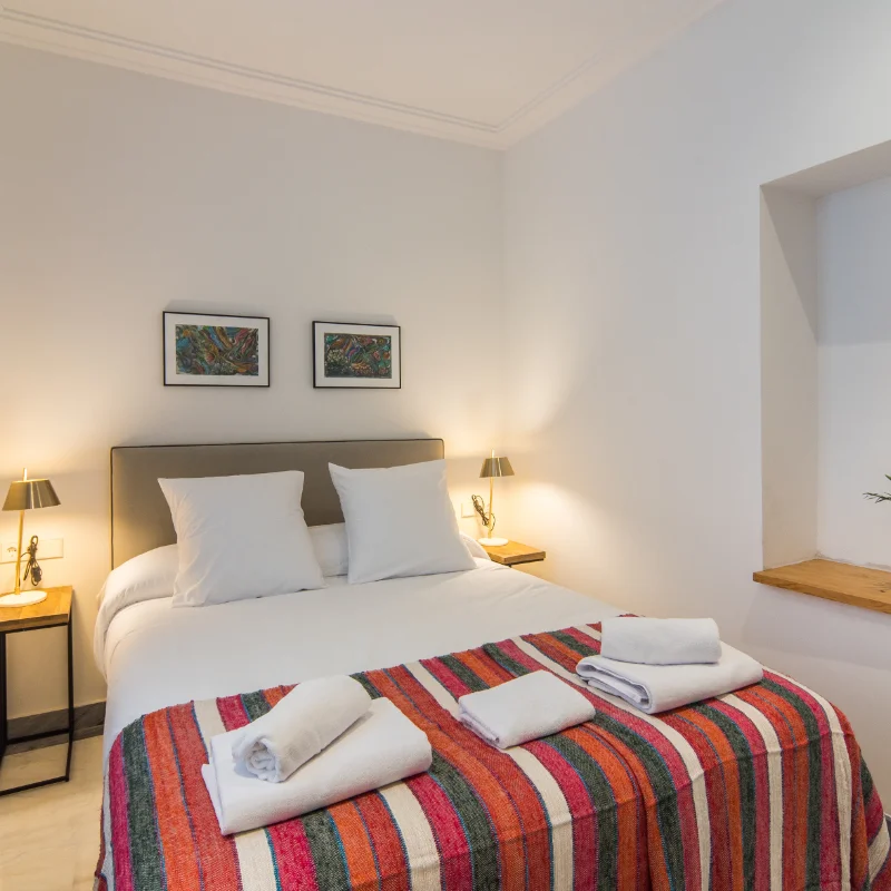 Cama doble con cobertor colorido y toallas en habitaci&oacute;n luminosa y moderna.