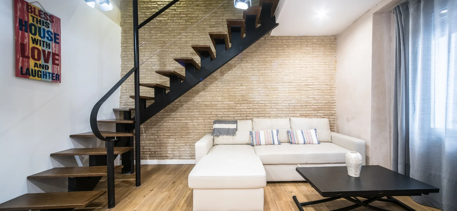 Escalera moderna junto a sof&aacute; beige, ladrillo expuesto y mesa negra.
