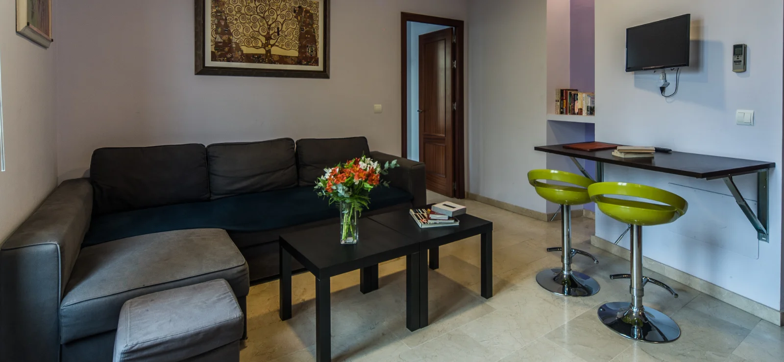 Sala de estar con sof&aacute;, mesa, flores, barra y taburetes verdes.