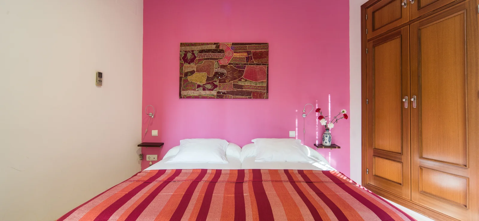 Cama con colcha rayada, pared rosa y armario de madera.