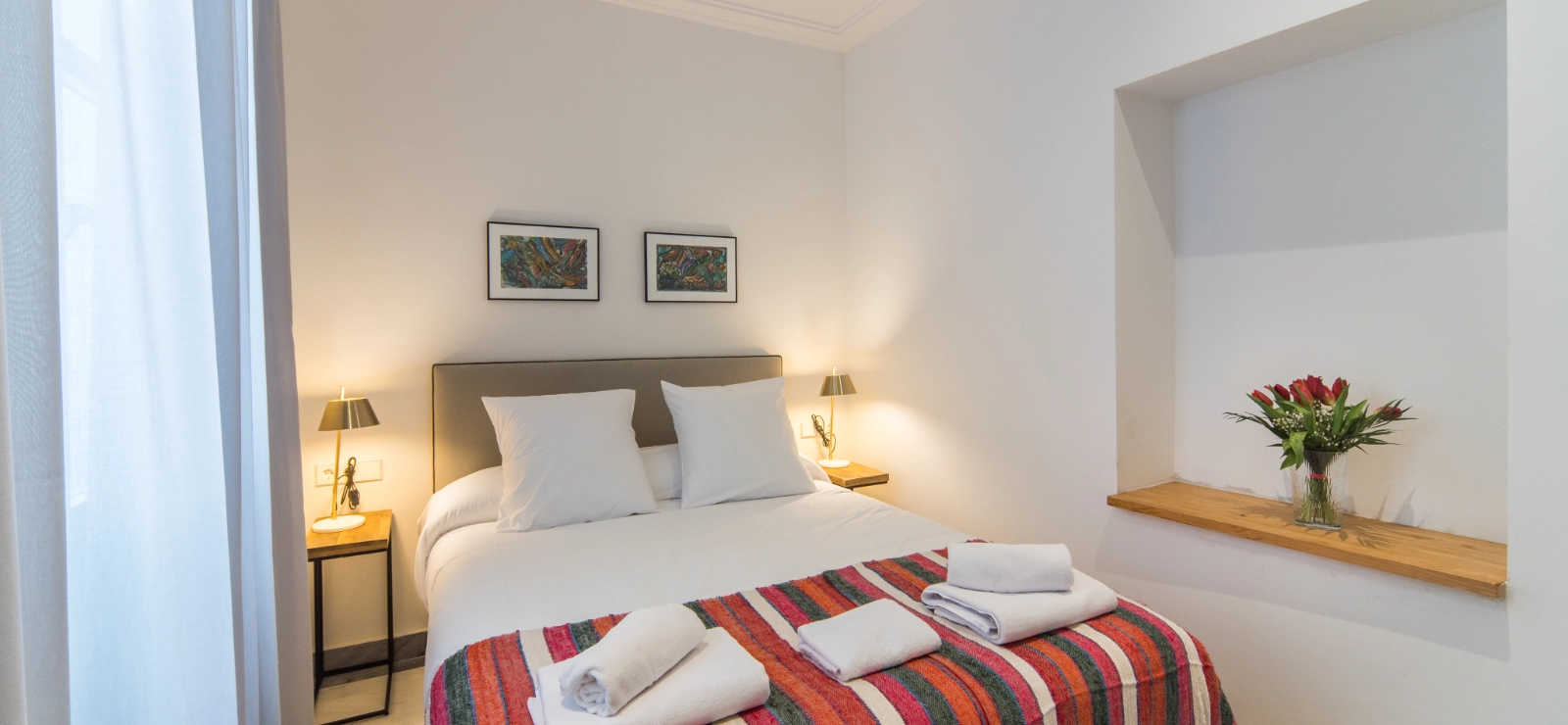 Habitaci&oacute;n luminosa con cama, colcha colorida y flores en un estante.