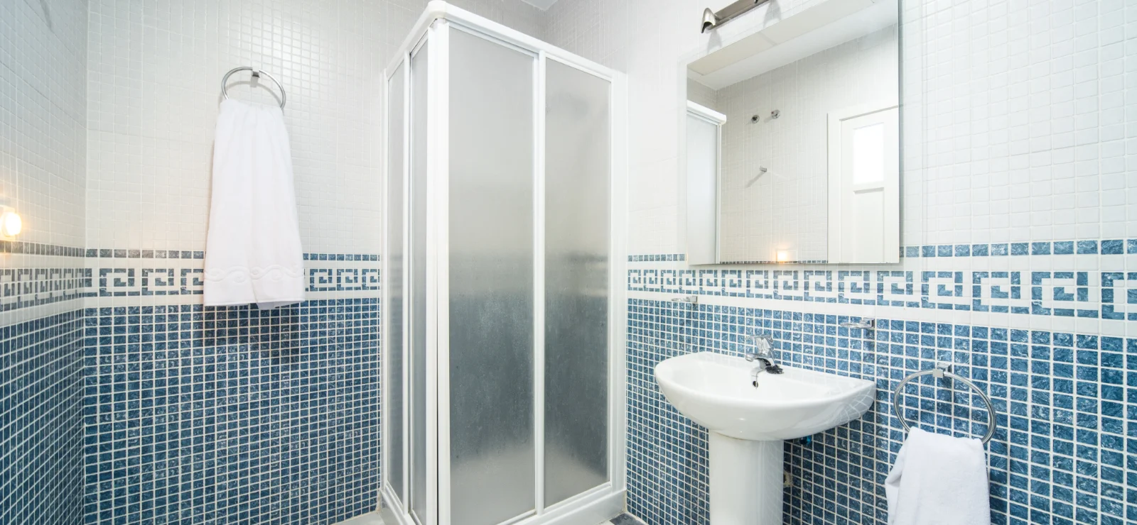 Ba&ntilde;o con azulejos azules, ducha, lavabo y toallas blancas.