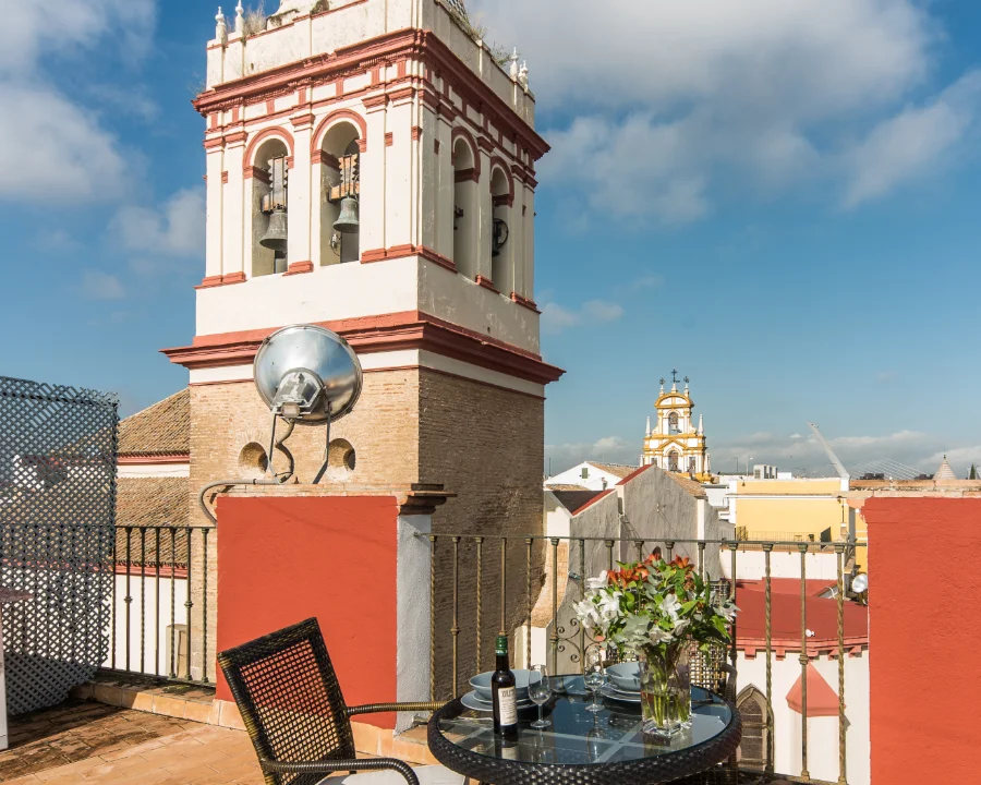 Terraza con mesa, sillas y flores, vista de campanario y cielo azul.