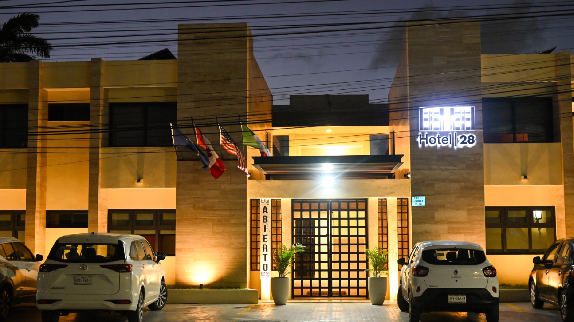 Fachada iluminada de un hotel con autos estacionados al frente.