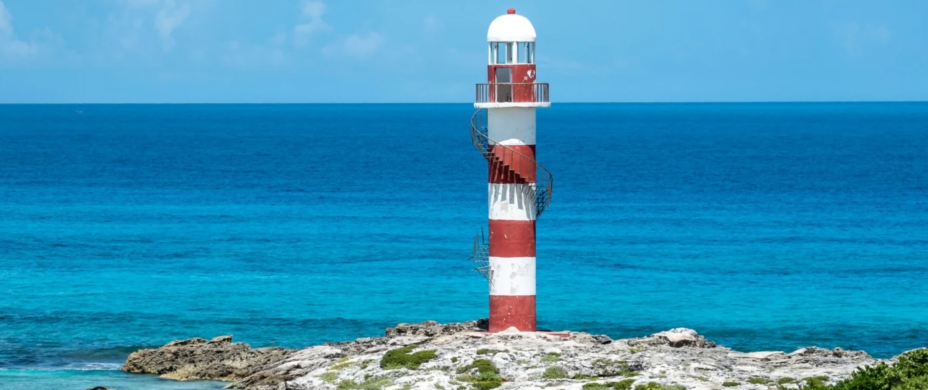 Faro a rayas en un acantilado frente a un mar turquesa.