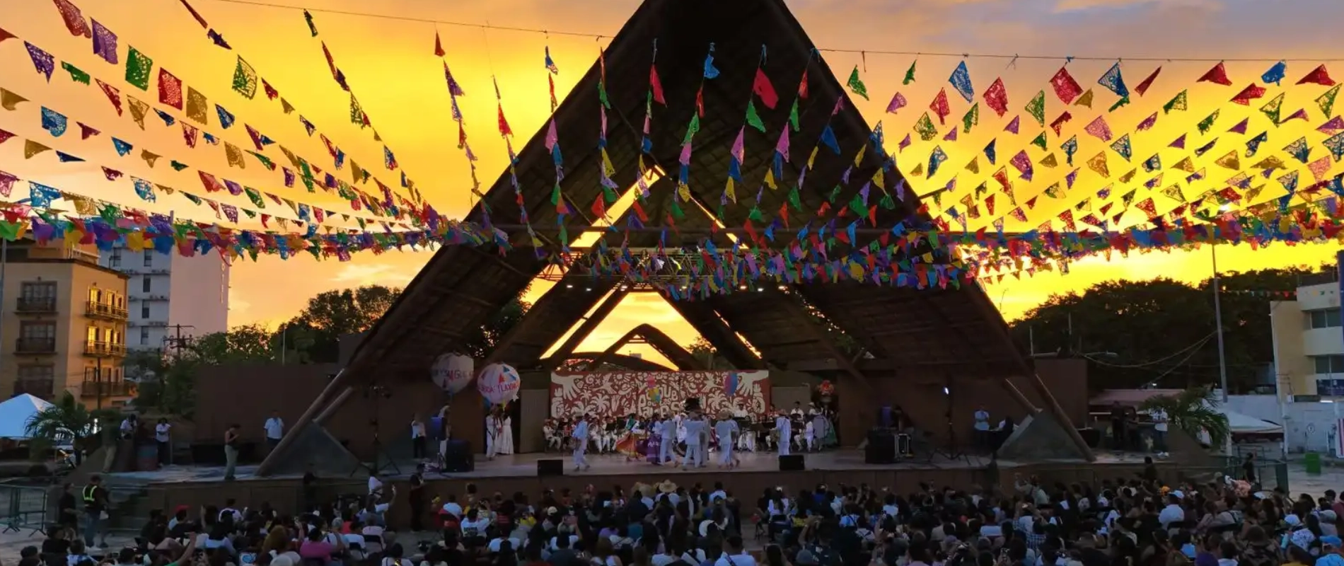 Festival al atardecer bajo papel picado colorido y escenario con p&uacute;blico.