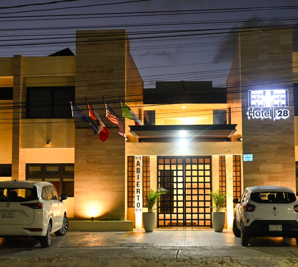 Fachada iluminada de un hotel, con autos y banderas.