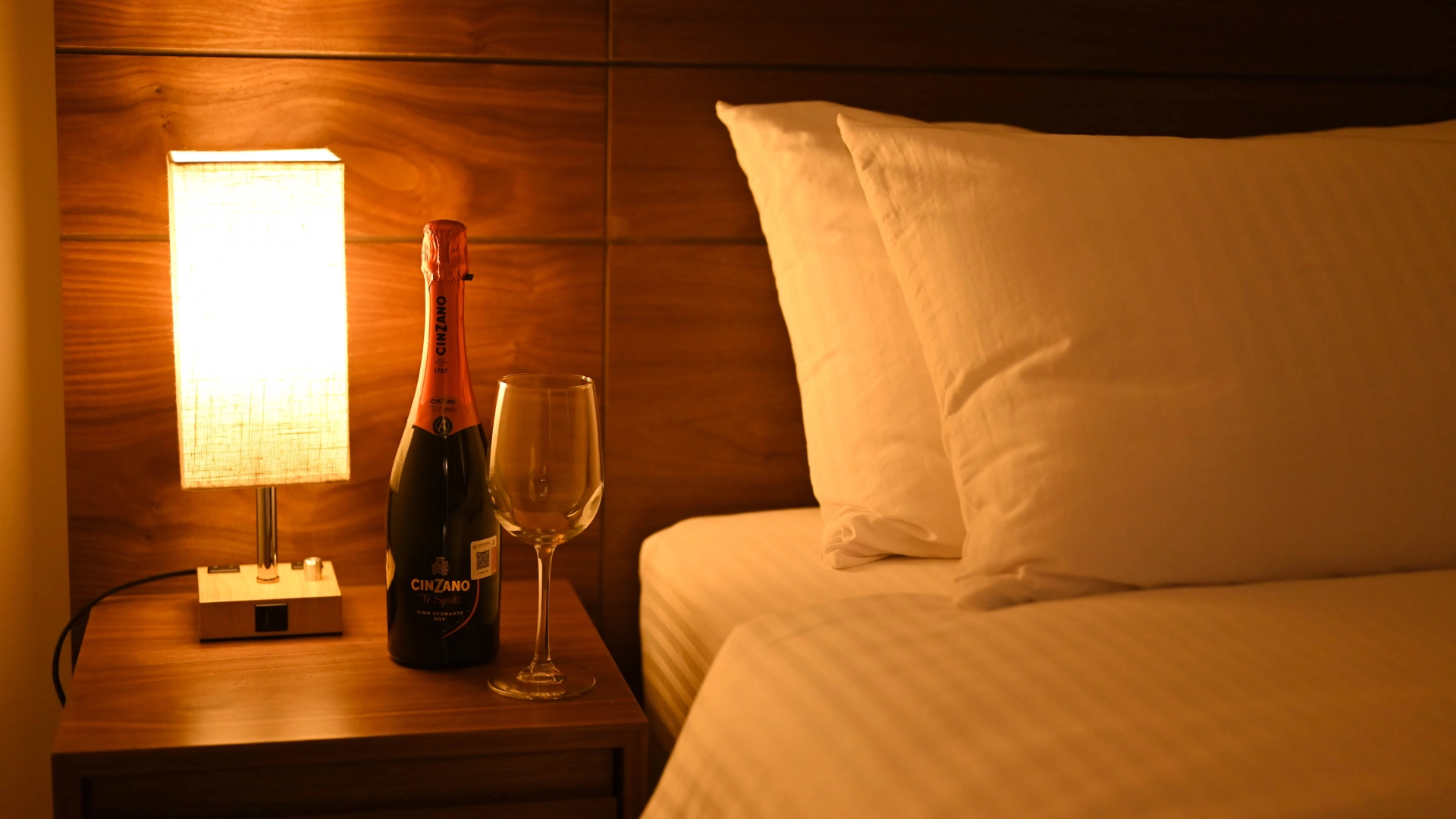L&aacute;mpara, botella y copa junto a una cama con almohadas blancas.