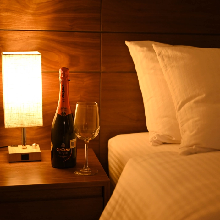 Botella y copa junto a l&aacute;mpara y cama con edred&oacute;n blanco en habitaci&oacute;n c&aacute;lida.