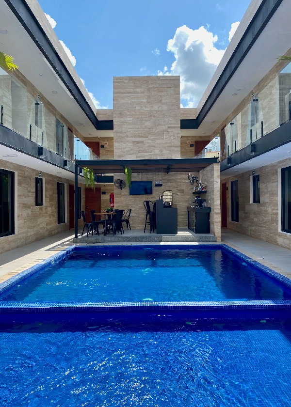 Patio moderno con piscina azul rodeada de paredes beige, bar y sillas negras.