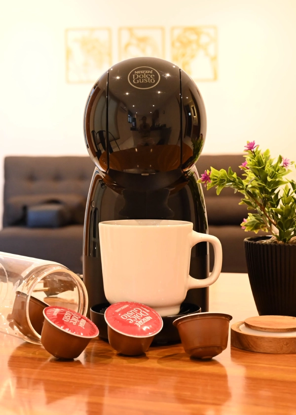 Cafetera Dolce Gusto con taza, c&aacute;psulas y planta en mesa de madera.