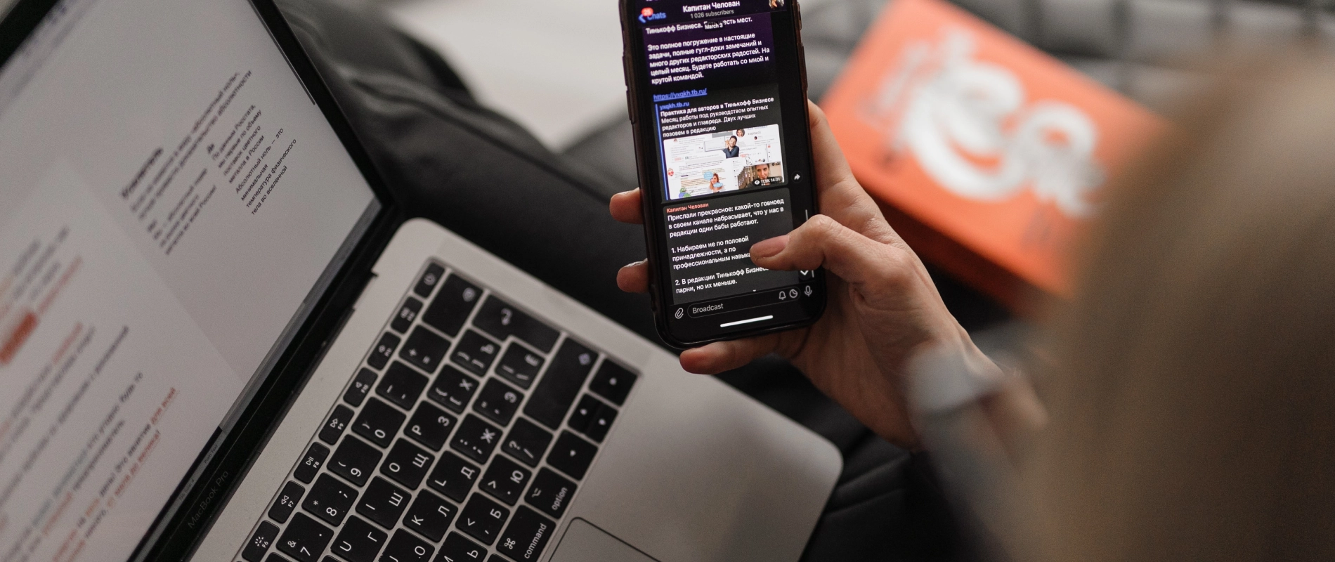 Mano sosteniendo un tel&eacute;fono m&oacute;vil frente a una laptop con texto en pantalla.