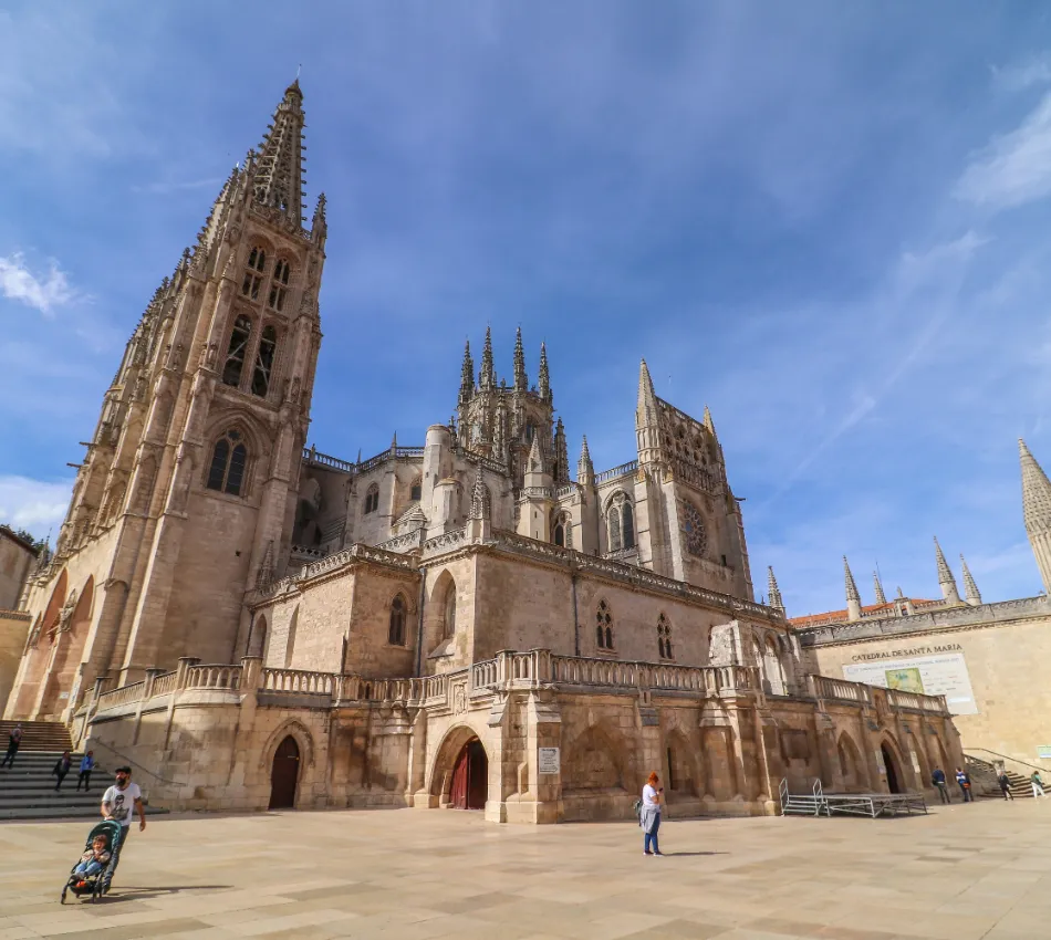 Catedral g&oacute;tica con torres y plaza, cielo despejado. Personas caminando alrededor.