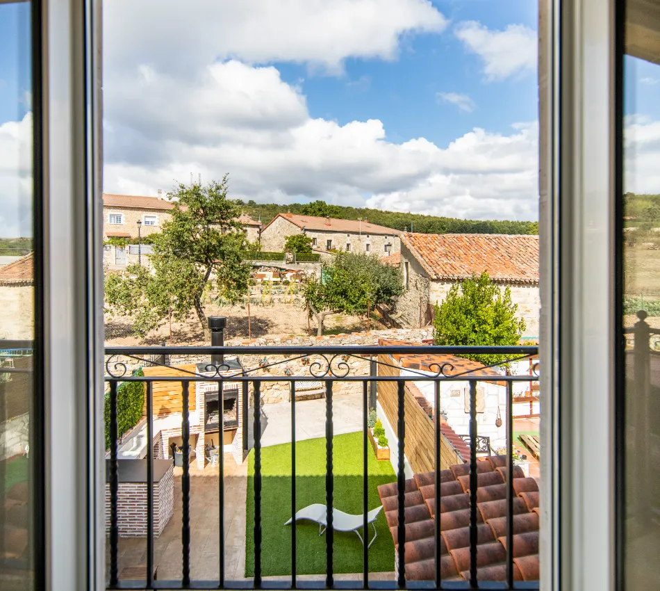 Vista desde una ventana hacia un jard&iacute;n con c&eacute;sped y tumbona, rodeado de casas de campo.