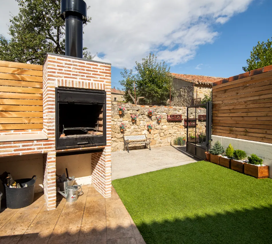 Patio con barbacoa de ladrillo, banco, c&eacute;sped artificial y pared con macetas de flores.