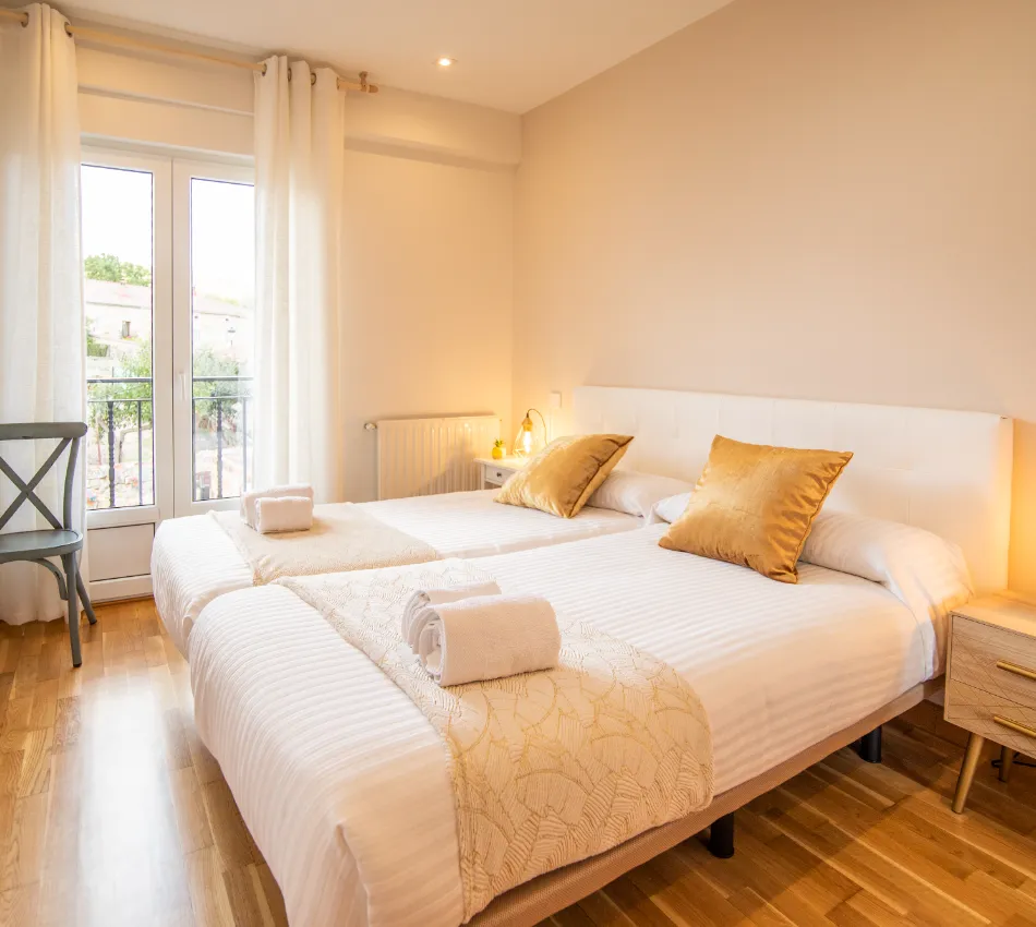 Habitaci&oacute;n luminosa con dos camas, cojines dorados y ventanal con cortinas blancas.