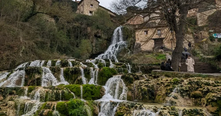 Cascada fluye entre musgo y piedras; casas de piedra y &aacute;rboles al fondo. Personas observan.