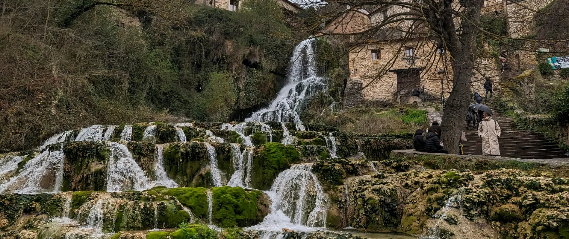 Cascada rodeada de musgo y vegetaci&oacute;n junto a un edificio de piedra y escaleras con visitantes.