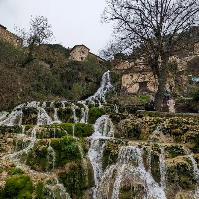Cascada en un pueblo con edificios de piedra y personas en el paisaje. &Aacute;rboles sin hojas.