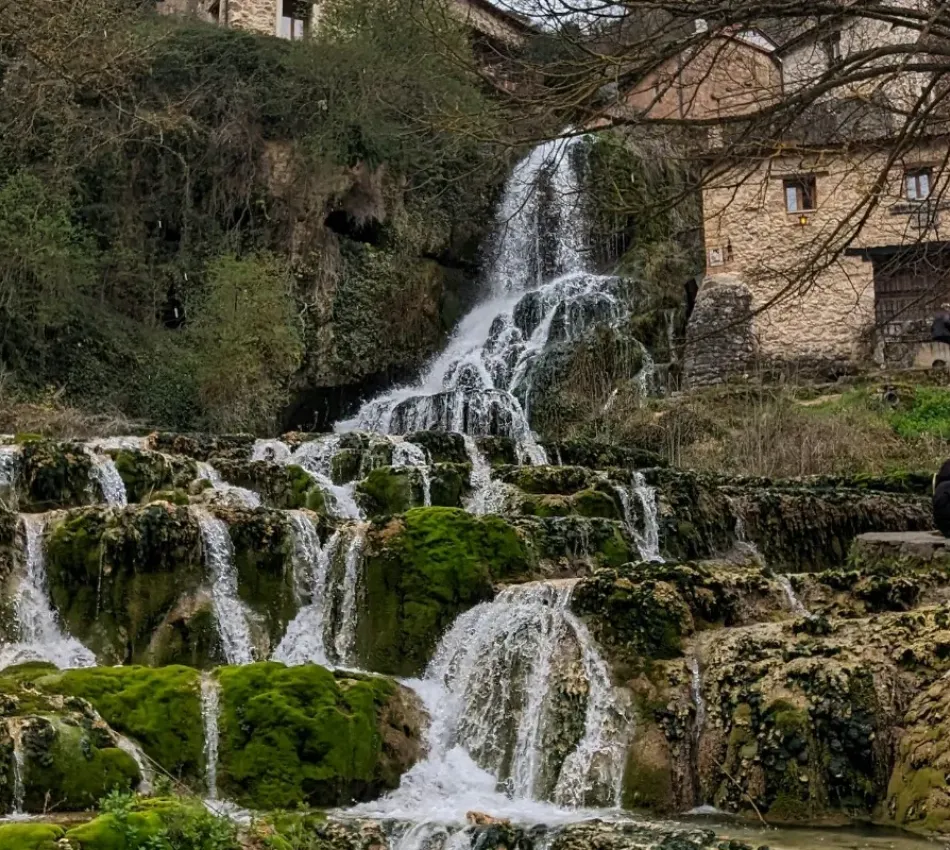 Cascada de agua fluye sobre rocas cubiertas de musgo, con una casa de piedra al fondo.