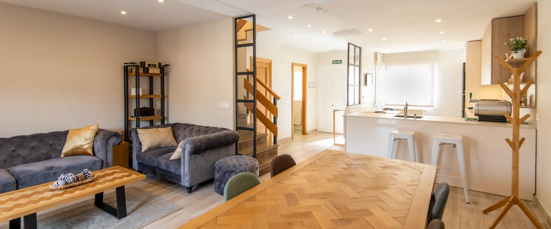 Sala moderna con sof&aacute;s grises, mesa de madera, y cocina abierta con barra y taburetes blancos.