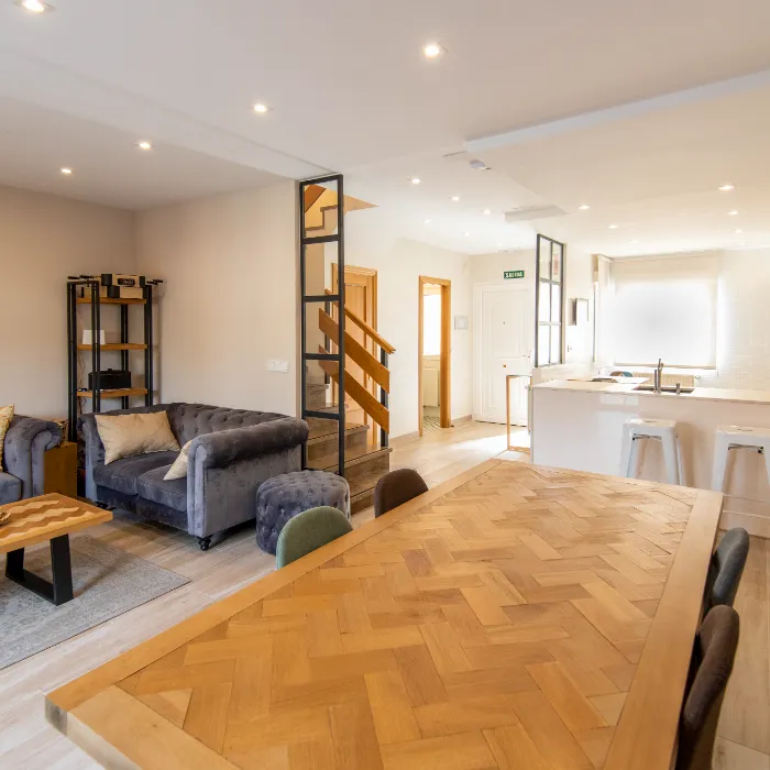 Sala luminosa con sof&aacute;, mesa de madera grande y cocina integrada con taburetes.