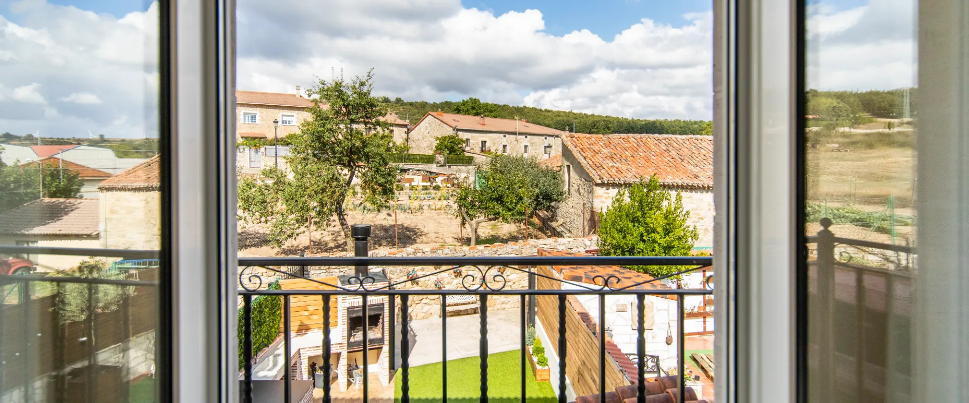 Ventana con vista a un patio y casas de techo de tejas, &aacute;rboles y cielo con nubes.