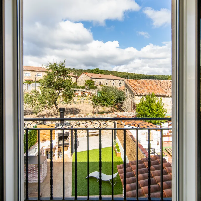 Vista desde una ventana hacia un jard&iacute;n con c&eacute;sped y tumbona, casas de piedra al fondo.