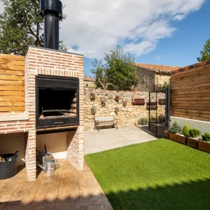 Patio con c&eacute;sped artificial, parrilla de ladrillo, y banco junto a un muro de piedra decorado.