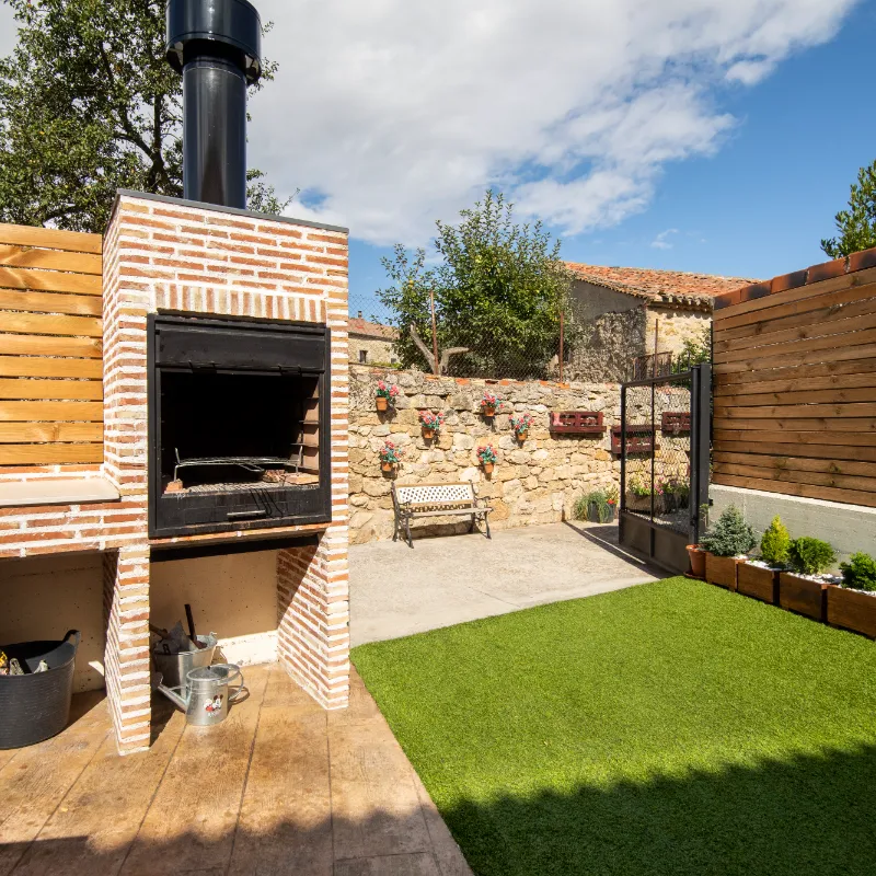 Patio con c&eacute;sped artificial, parrilla de ladrillo, y banco junto a un muro de piedra decorado.