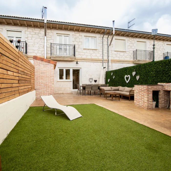 Patio con c&eacute;sped, tumbona, sof&aacute;, mesa y paredes de ladrillo; decoraci&oacute;n de corazones en arbustos.