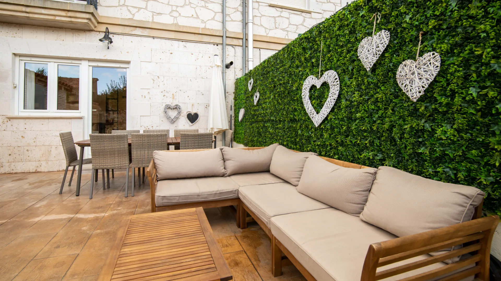 Terraza con sof&aacute; beige, mesa de madera, sillas de mimbre y pared verde decorada con corazones.