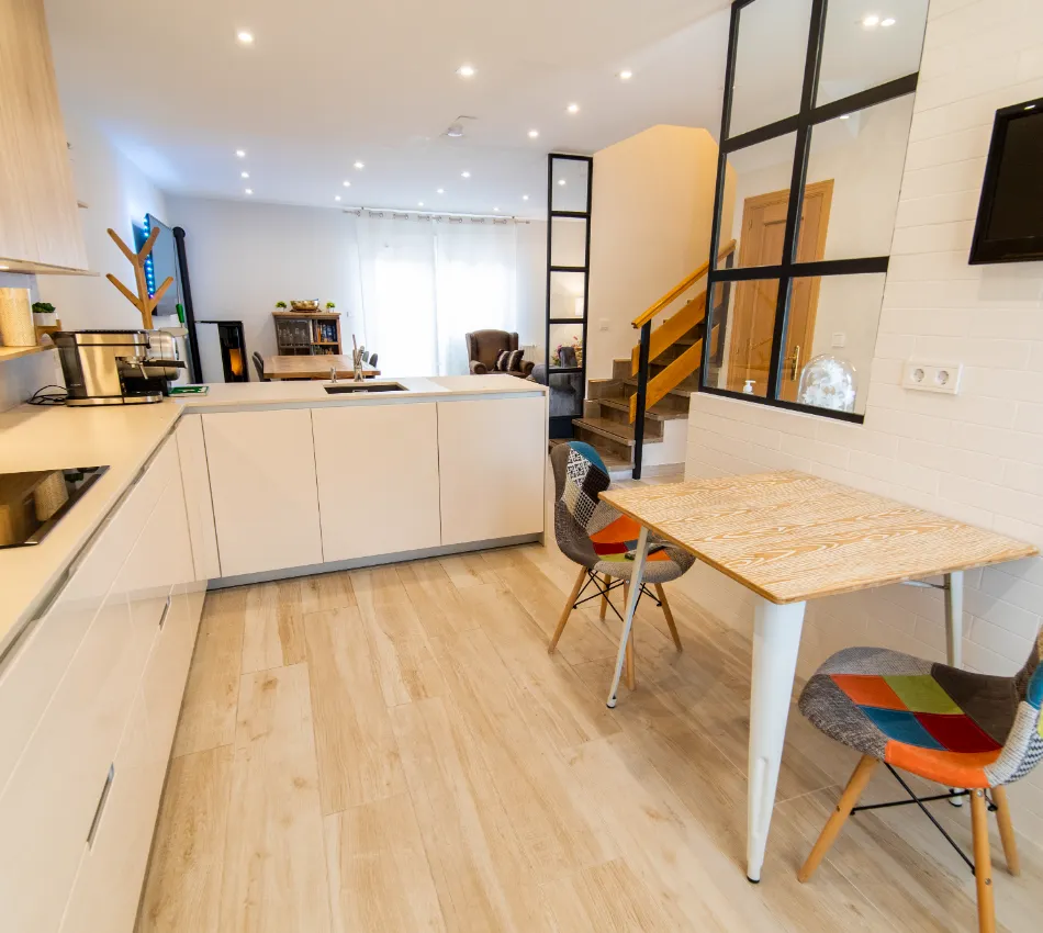 Cocina moderna con mesa de madera, sillas de colores y escalera en el fondo.