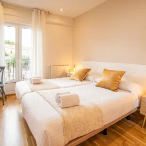 Habitaci&oacute;n con dos camas, cojines dorados y ventanal con cortinas blancas. Decoraci&oacute;n acogedora.