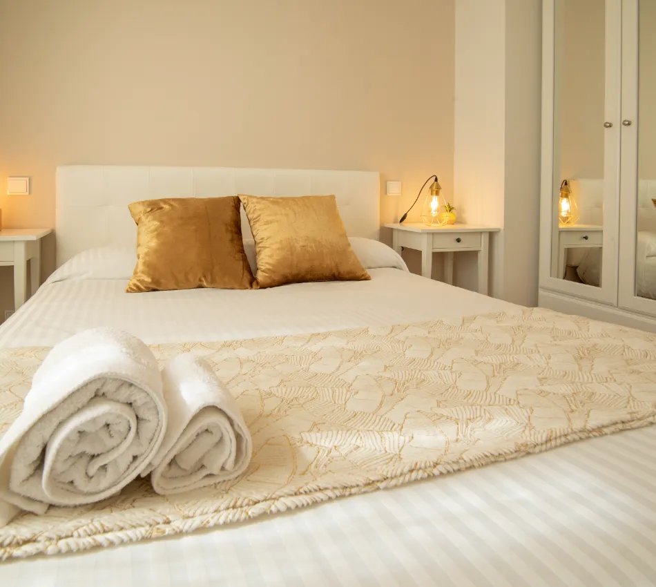 Cama elegante con cojines dorados, l&aacute;mparas y toallas enrolladas sobre un cubrecama beige.